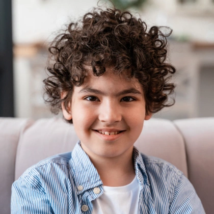 Smiling young boy with curly brown hair in dental office setting, wearing blue striped shirt.