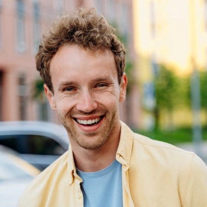 Smiling man in yellow shirt standing outdoors near parked car and buildings.