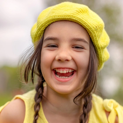 Young girl smiling brightly wearing yellow beanie and braids outdoors.