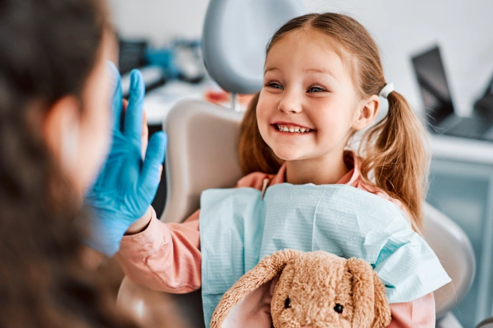 Young girl smiling at dentist during checkup, holding stuffed animal in dental office.