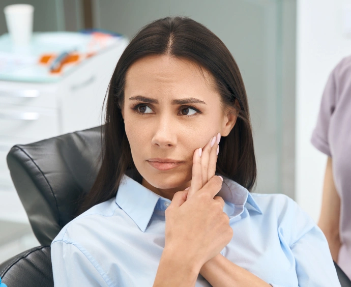 Woman in light blue shirt sitting at desk with thoughtful expression, hand to face, in dental office setting.