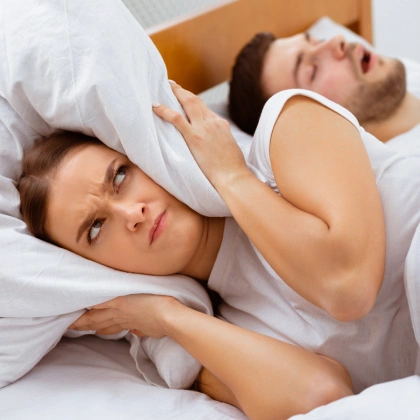 Woman covering ears in bed while man snores beside her.