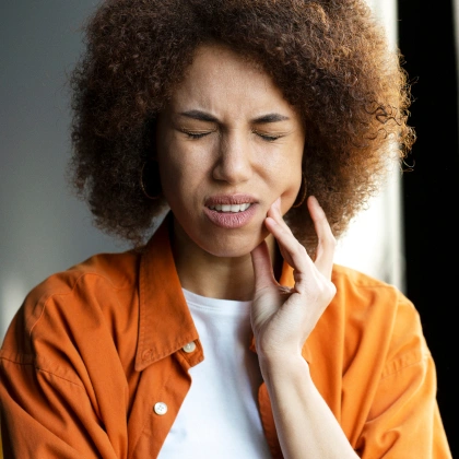 Woman in orange shirt touching her cheek, appearing to have tooth or jaw discomfort, neutral background.