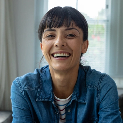 Smiling woman in denim shirt sitting indoors near window, dental practice portrait.