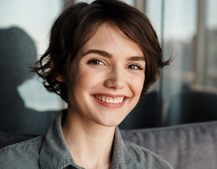 Smiling woman with short dark hair in modern office setting with city skyline visible behind her.