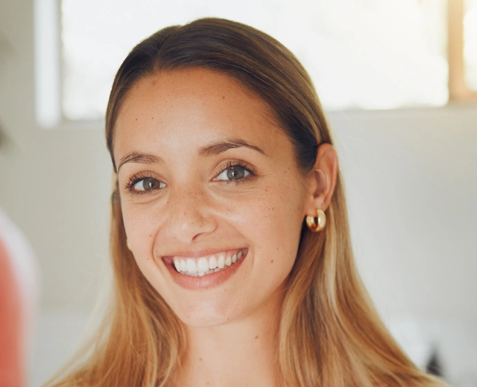Young woman smiling at camera in bright dental office setting, wearing pearl earrings.