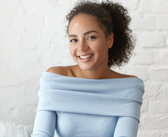Smiling woman wearing light blue off-shoulder top against white brick wall background.