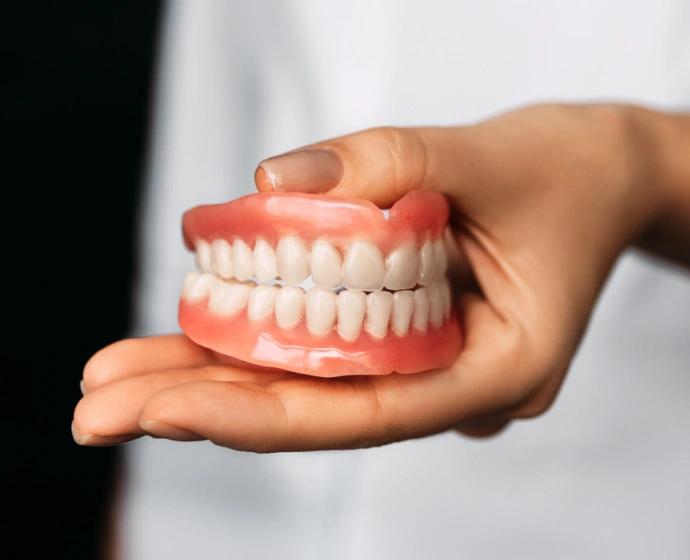 Hand holding a full set of dentures with pink gums and white teeth against white background.