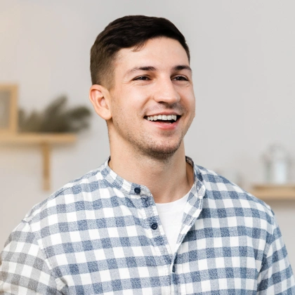 Young man smiling in casual checkered shirt against blurred dental office background.