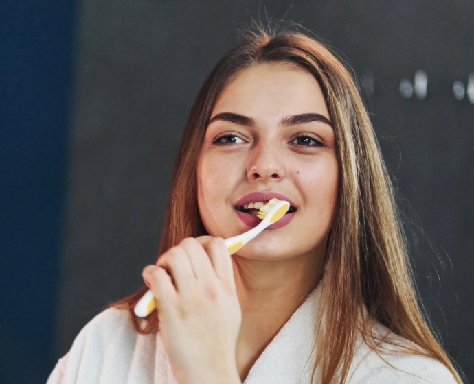 Young woman brushing teeth in bathroom with white robe, smiling at camera.