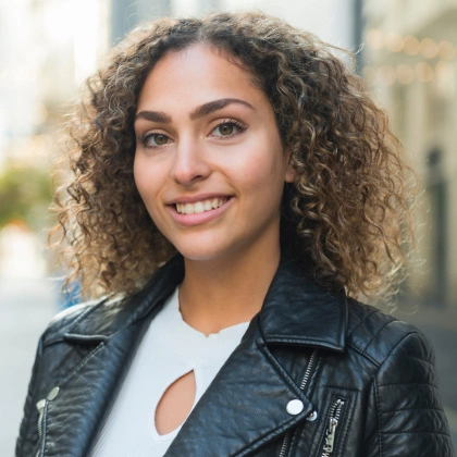 Professional headshot of smiling woman with curly hair wearing black leather jacket outdoors.