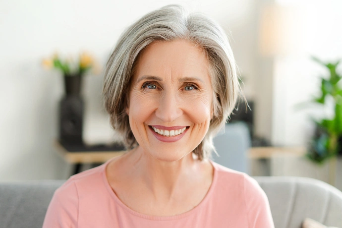 Smiling woman with gray hair wearing pink shirt in modern dental office setting.