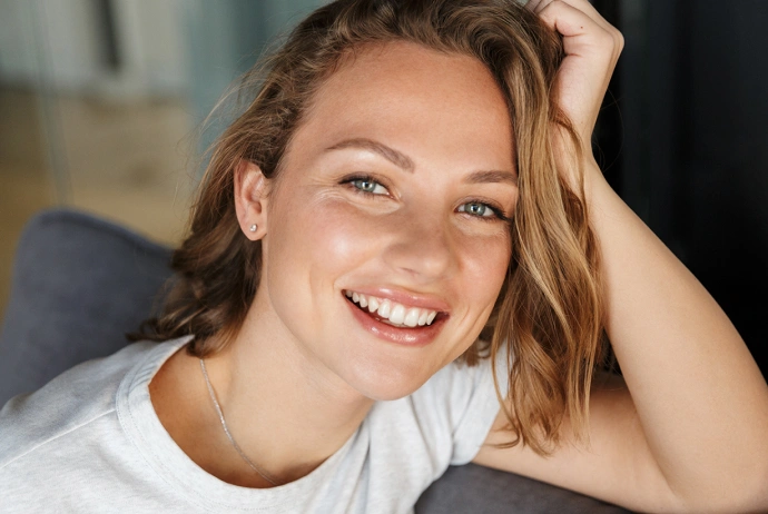 Young woman smiling at camera, hand in hair, wearing white shirt, casual indoor setting.