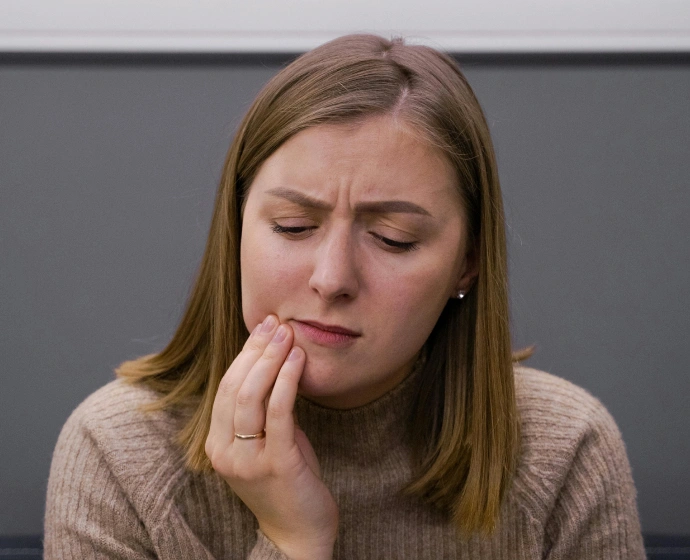 Woman with concerned expression touching her mouth, suggesting dental discomfort or worry against gray background.