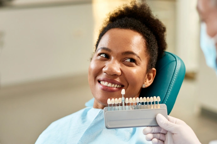 Patient smiling in dental chair while dentist holds tooth shade guide for color matching.
