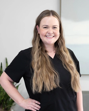 Professional woman with long brown hair smiling at camera in dental office setting, wearing black shirt.