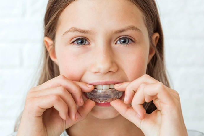 Young girl inserting clear orthodontic aligner into mouth against white brick background.