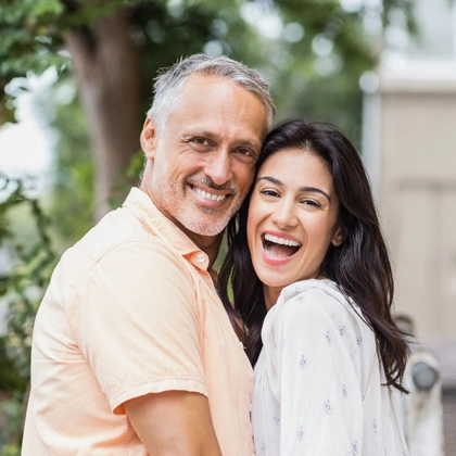 Smiling mature man and woman hugging outdoors in casual clothing near greenery.