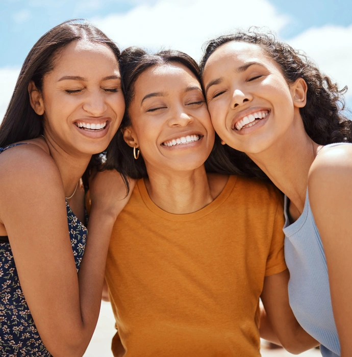 Three smiling women posing together outdoors against blue sky background.