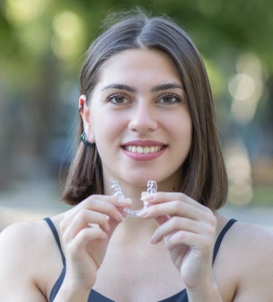 Young woman smiling outdoors, holding clear dental aligner in front of blurred green background.