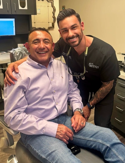 Two men smiling at camera in dental office setting with equipment visible in background.