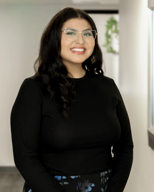 Woman wearing glasses and black sweater smiling in dental office hallway setting.