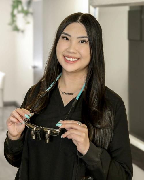 Smiling dental professional holding dental tools in modern clinic setting wearing black scrubs.