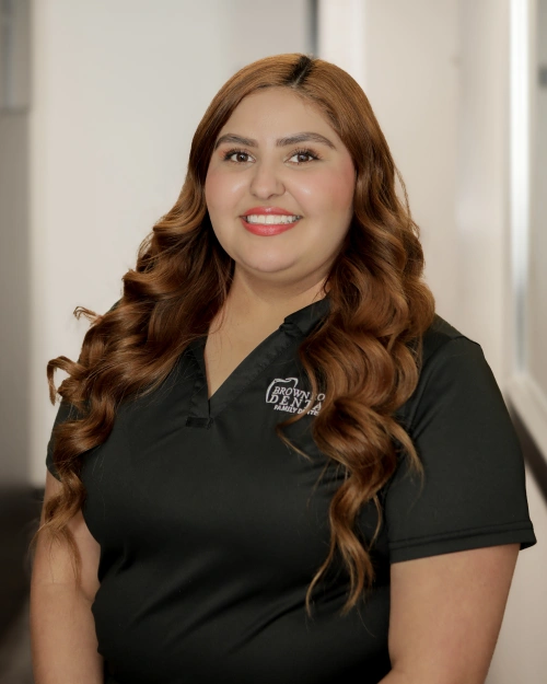 Female dental professional with long brown wavy hair wearing black Brown Road Dental polo shirt, smiling at camera in clinical setting.